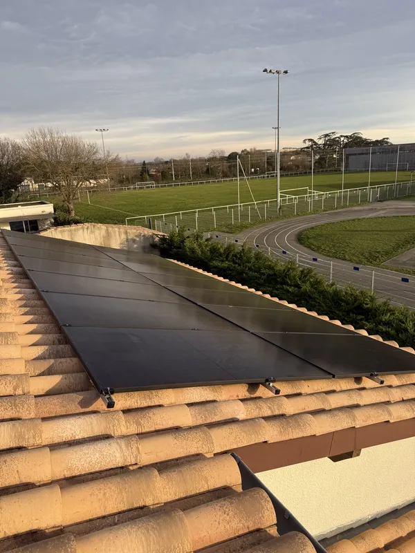 Panneaux photovoltaïques noirs sur une toiture en tuiles canal à Vernet, avec le terrain et la piste d'un stade derrière, janvier 2026